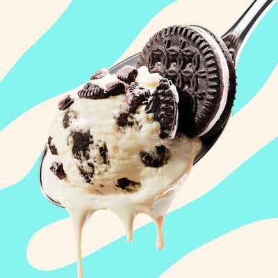 Pint of N!CK'S Cookies & Cream light ice cream. The label highlights 'no added sugar' and '330 calories/pint'. A spoon holds a scoop of vanilla ice cream with dark cookie pieces.