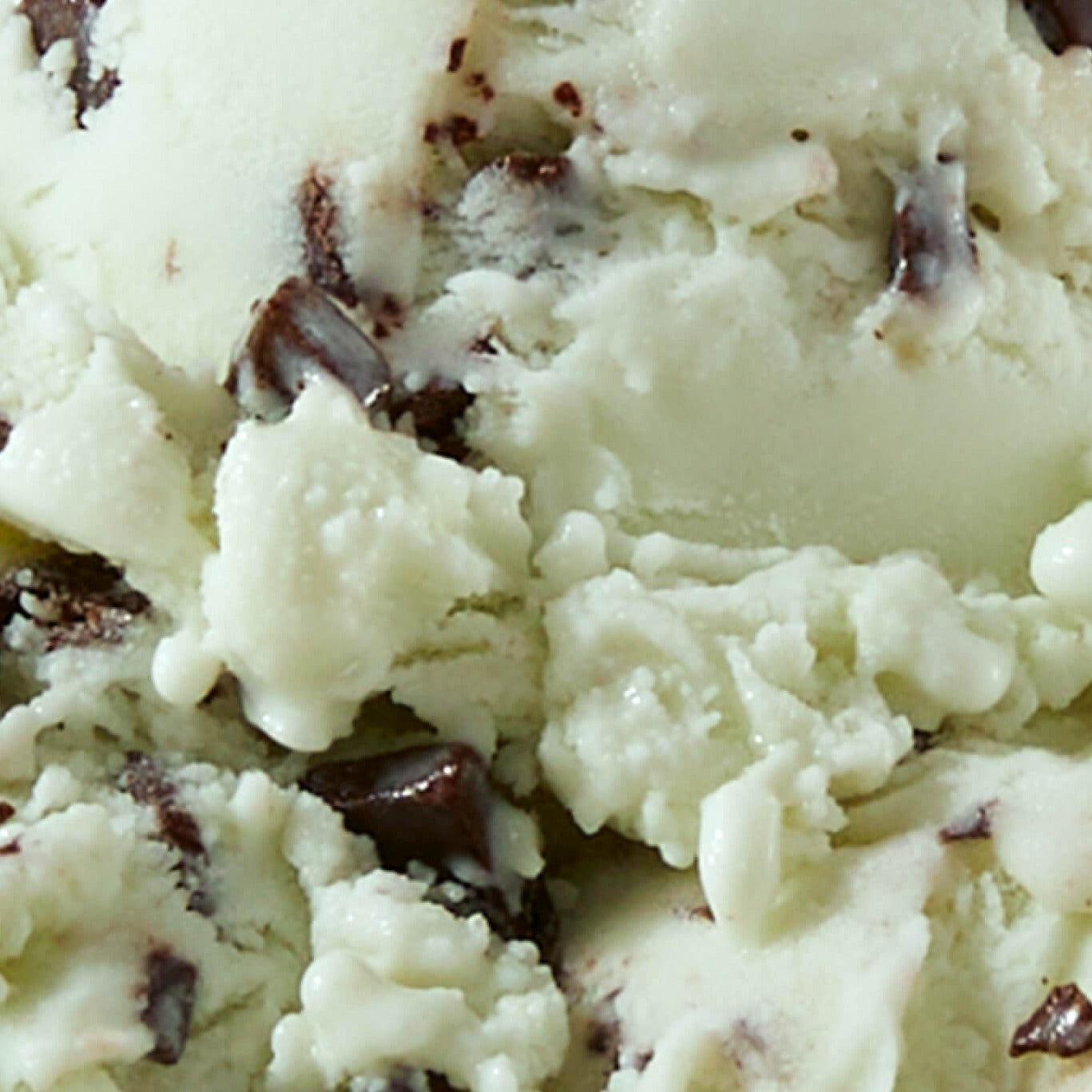 Extreme close-up texture shot of N!CK'S Mint Chocolate Chip light ice cream, showing pale mint green ice cream with visible chunks of dark chocolate chips.