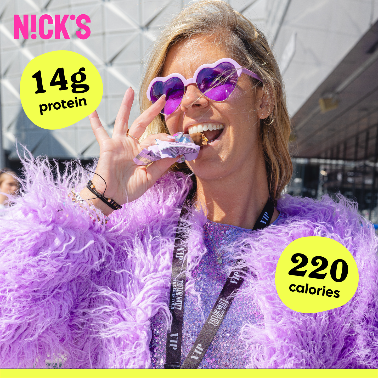 Smiling woman wearing purple faux fur and heart-shaped sunglasses, eating a N!CK'S Almond Chocolate Protein Bar. Text highlights '14g protein' and '220 calories'.